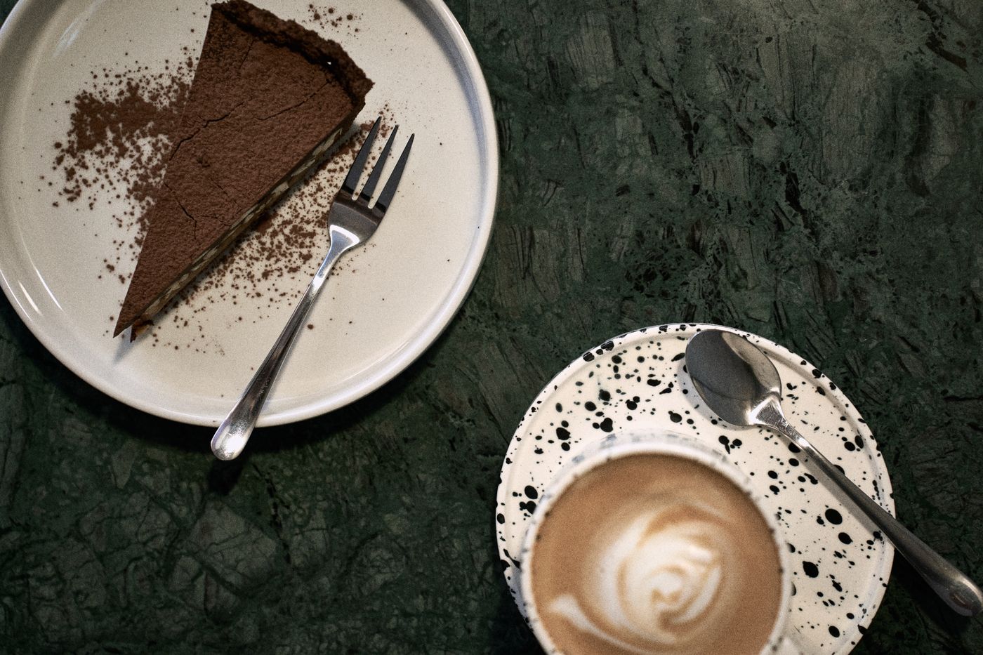 Tarta czekoladowa posypana kakao i flat white z wzorem z pianki w nakrapianej filiżance, na zielonym marmurowym stoliku w Bruk Cafe